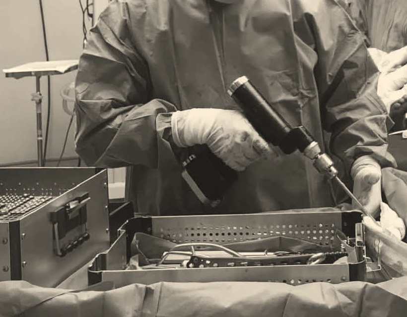 An operating-room back table: a powered driver in gloved hands beside an open instrument tray.