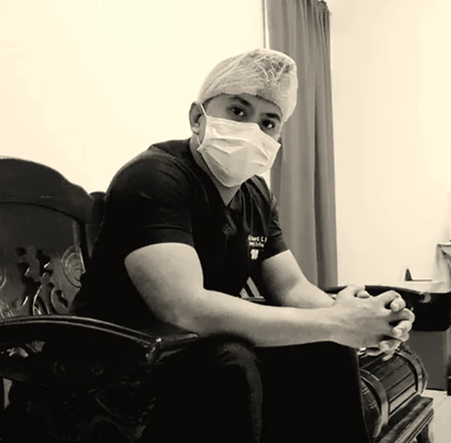 Richard Cas in scrubs, cap and mask, sitting outside theatre with his hands clasped.